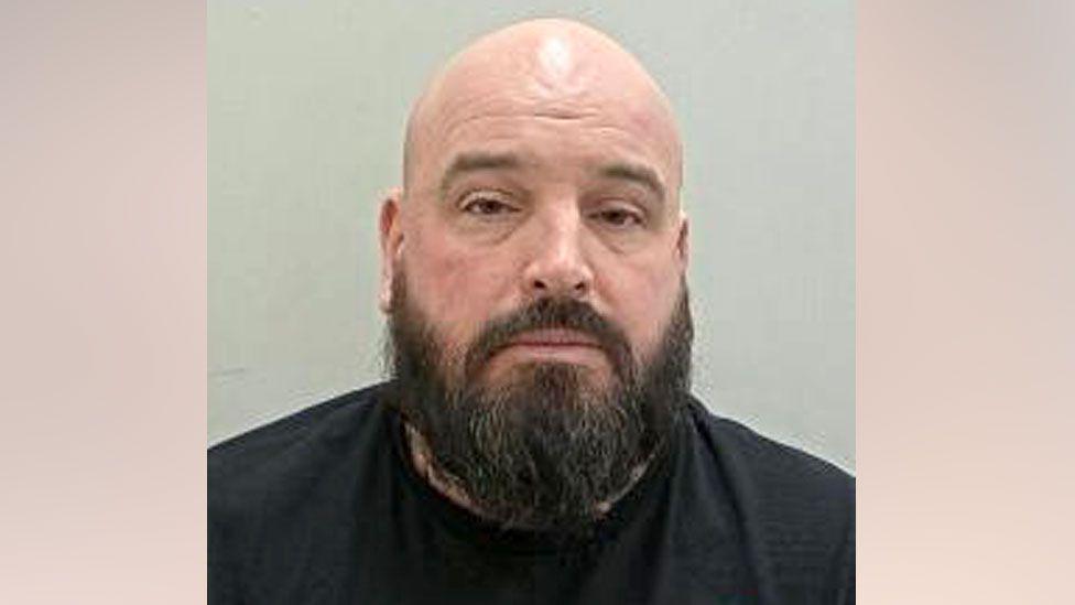 A police mugshot of Neil Platt, who has a bald head and a thick black beard.