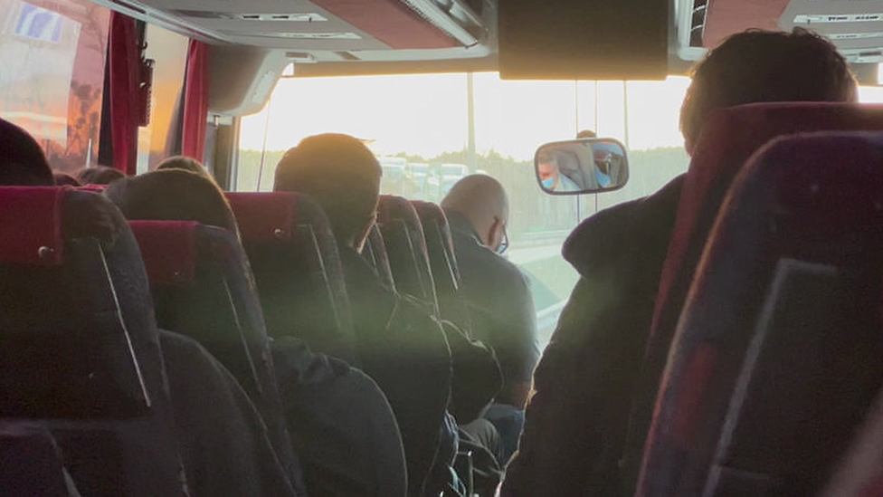 A group of people, seen from behind, sitting on a coach as it travels down a main road. The vehicle appears to be full with passengers, and bright sunlight fills the horizon in the distance.