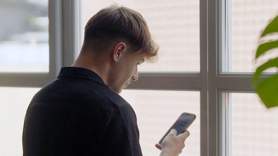 Cai, now aged 18, looking at his phone as he faces a large window