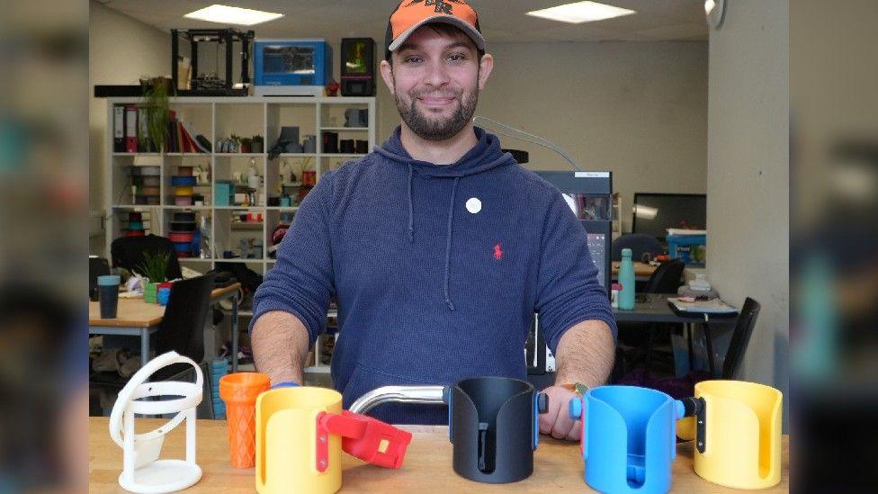 Michael Rose in a dark blue hoodie and orange baseball cap, with some of his products - brightly coloured wheelchair cup holders, and a large plastic ice cream cone