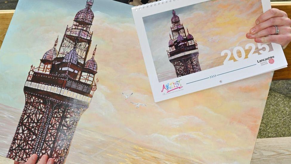 Kathryn Olley's winning painting features a close-up view of Blackpool Tower, set against a yellow and blue sky. Two seagulls flying above the sea reflects the same colours. In the top right of the picture we see a copy of the same painting on the front cover of a calendar for 2025.