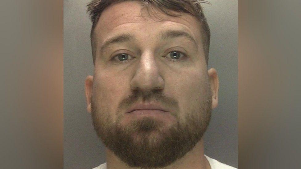 Dawid Dembler is pictured in a mugshot wearing a white t-shirt. He has light brown hair and a light brown beard.
