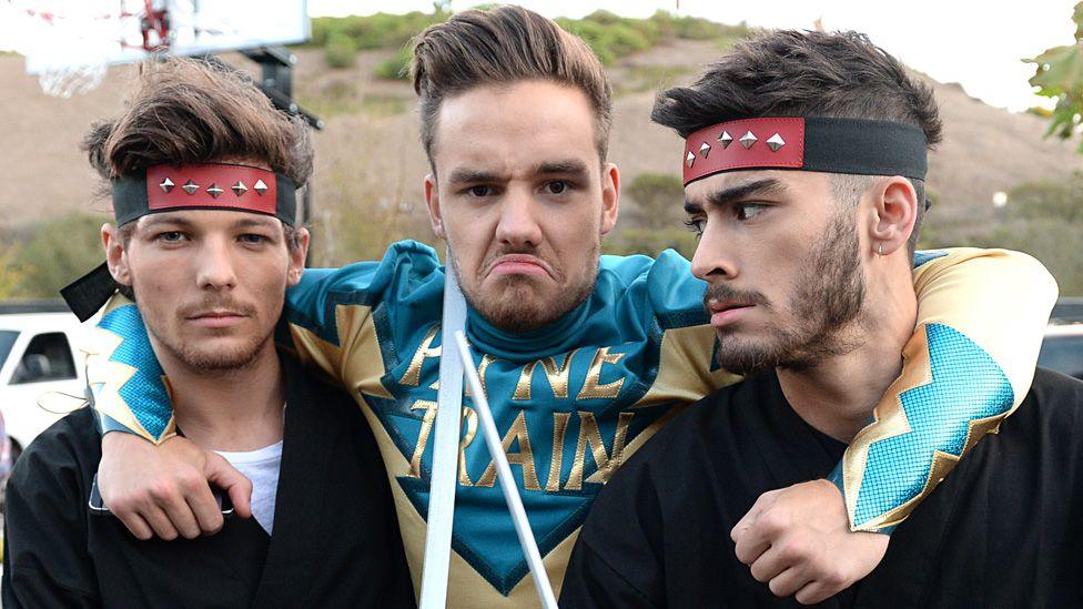 Liam Payne posing and pulling a face in a superhero "Payne Train" costume, with his arms around the necks of Louis Tomlinson and Zayne Malik, who are wearing superhero-style headbands