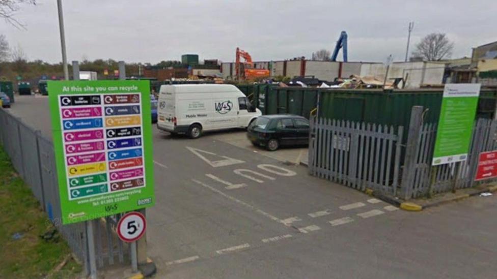 Entrance to Dorchester household recycling centre which is surrounded by steel fencing. Inside, cars are parked in a row in front of large open-top shipping containers.