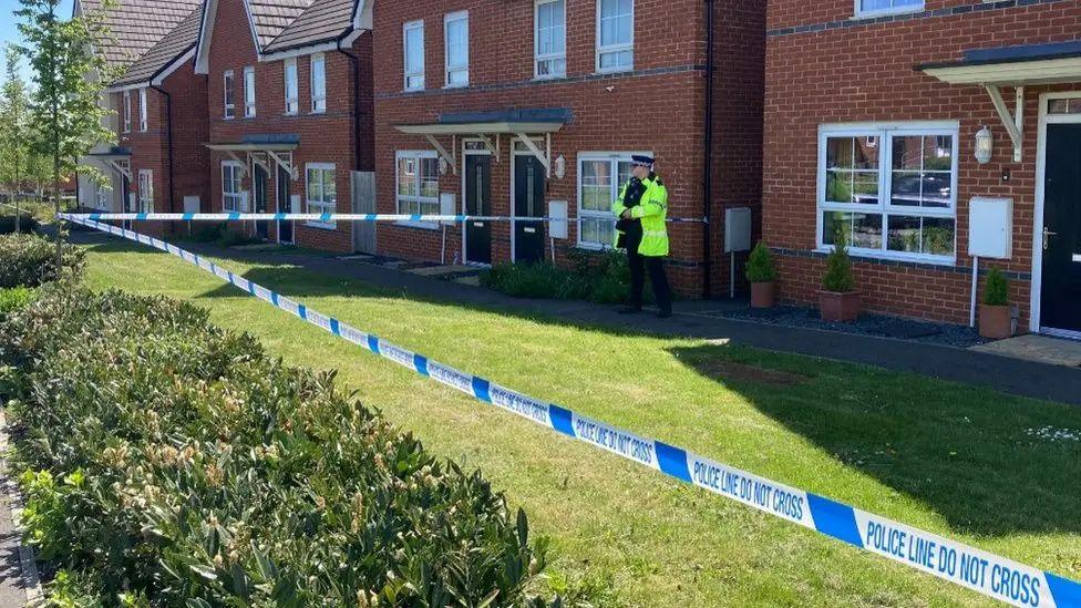 A police officer in a hi-vis jacket stands guard outside a line of semi-detached red-bricked houses.  There is a strip of lawn outside and blue and white police tape cordons off the house nearest the camera 