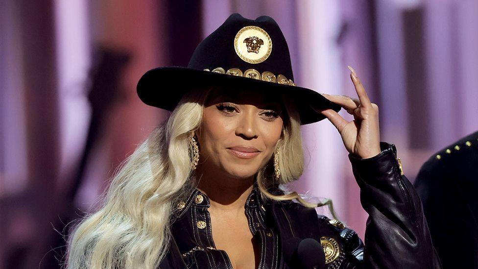 Beyoncé accepting an award on stage, wearing a black cowboy hat, using her left hand to hold the top of the hat. Her black outfit also has golden buttons.