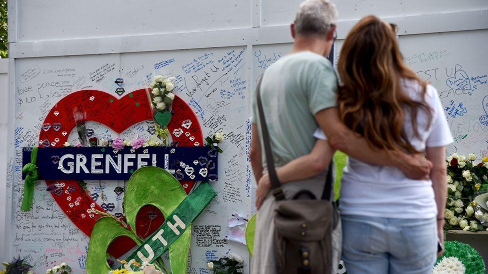 Two people hug in front of the Grenfell remembrance wall, with a heart in the background that says 'Grenfell' attached to the wall and messages people have written on the wall, in London 