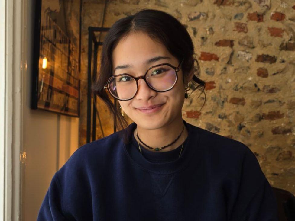 A woman in a blue jumper and dark glasses smiles in front of a brick wall