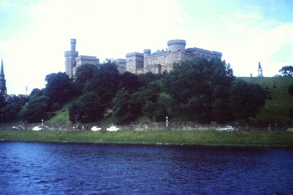 Castle  behind trees in front of a river, with old cars on a road in between.