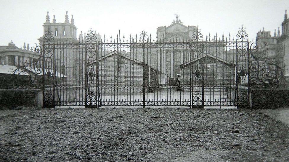 Black and white photograph of how Blenheim Palace looked during World War Two. There is a large metal fence in front of the huge grand building.