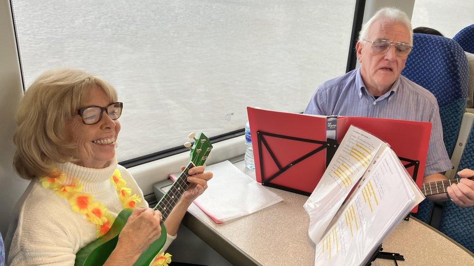 Two people play ukuleles while sitting on a train, with their music in front of them on stands