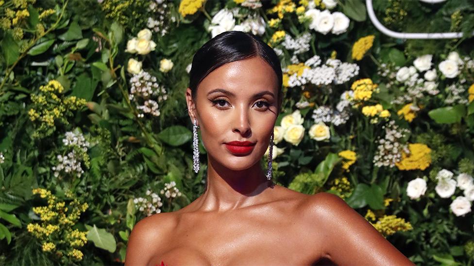 Maya Jama looking at the camera, wearing shiny earrings. The background is flowery with white, yellow flowers and green leaves.