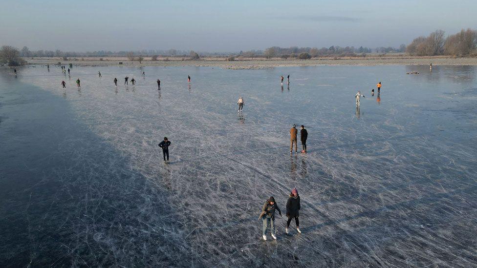 An aerial shot of dozens of people skating on a frozen field at Upware, Cambridgeshire. The blue ice is scored with white lines from the skates. In the distance can be seen the edge of the field and trees beneath a pale blue sky