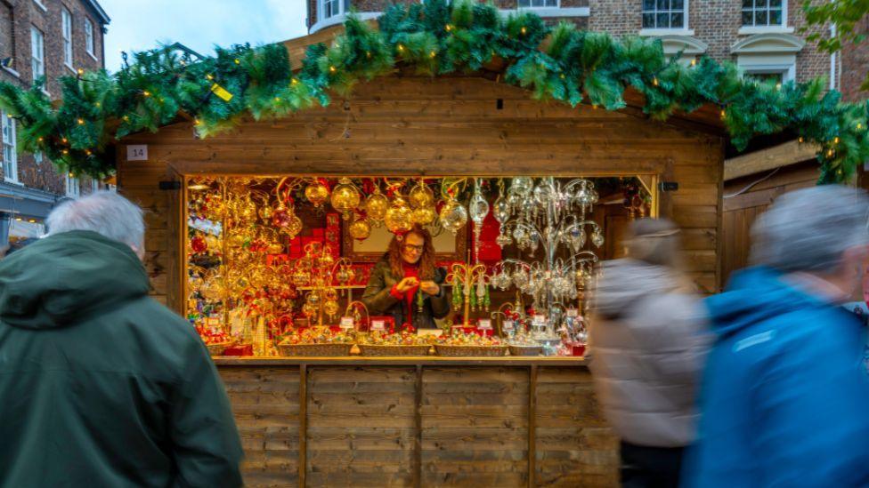 A wooden hut decorated with a Christmas theme with a woman displaying items for sale inside the hut as part of York's annual Christmas Market