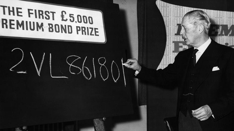 A black and white image of Prime Minister Harold MacMillan (1894-1986) writing the number of the first ever £5,000 Premium Bond prize on a chalkboard  at the National Savings headquarters in London.