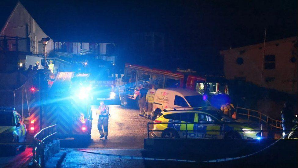 Police cars, fire engines and emergency services teams on a slipway - it is dark and blue flashing lights light up the area a building can be seen behind.