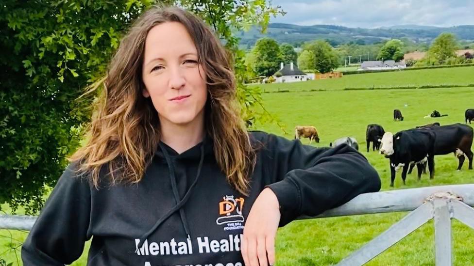 Menyw gyda gwallt hir brown yn sefyll tu fas yn siwmper sy'n dweud 'mental health' 