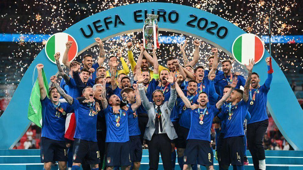 UEFA Euro 2020 Italy team celebrating the win, holding the trophy with sparkles from pyrotechnics behind them