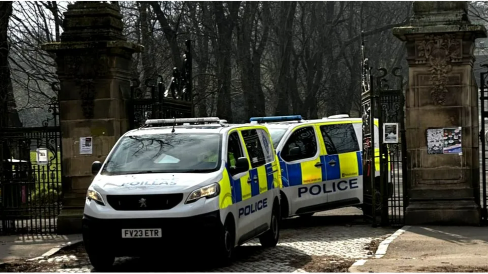 Two police vans parked between two large black iron gates and a wall- the entrance to a park