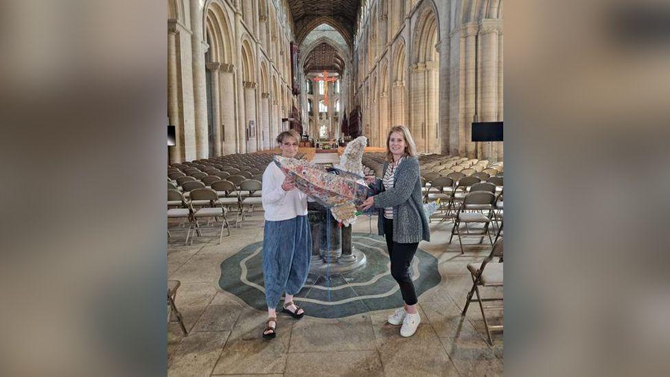 Two women holding the recycled shark model in the nave of Peterborough Cathedral. One is blonde and is wearing a grey cardigan and black trousers, while the other has short hair and is wearing glasses, a white top and baggy navy trousers.