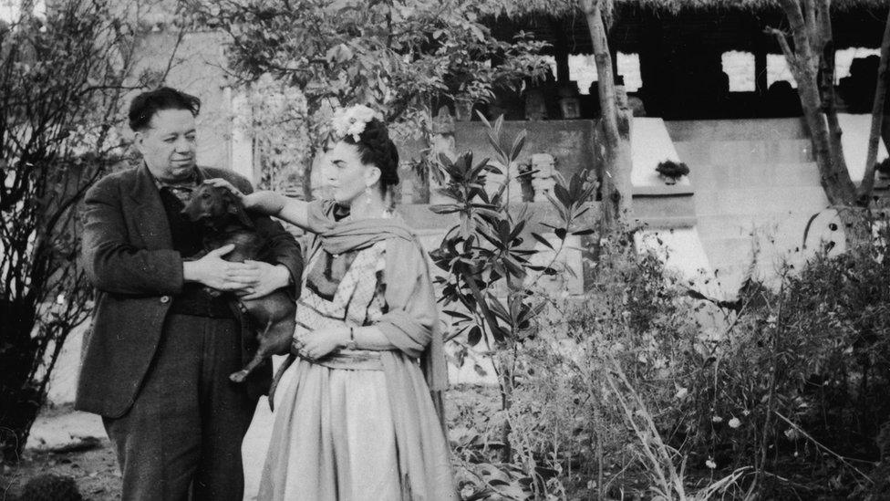 Mexican painters Frida Kahlo (1907 - 1954) and Diego Rivera (1886 - 1957) stand together with a pet dog in front of thatched-roof hut which houses a number of archaeological artefacts, Mexico City, Mexico, 1940s