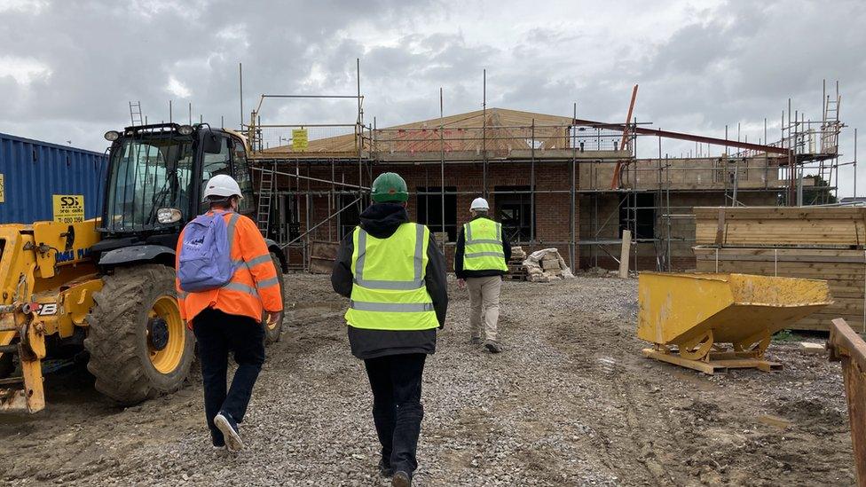 Three people in high vis and hard hats walking past a digger towards a construction site with a partially-done building.