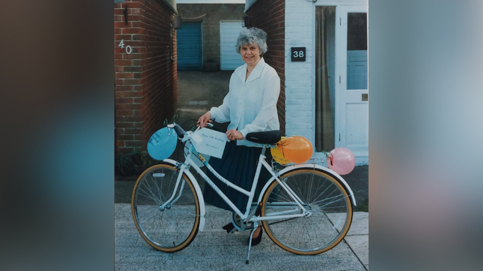 An archive image of Joan, who has grey hair and is wearing a white blouse, holding the bike - which has balloons tied to it.