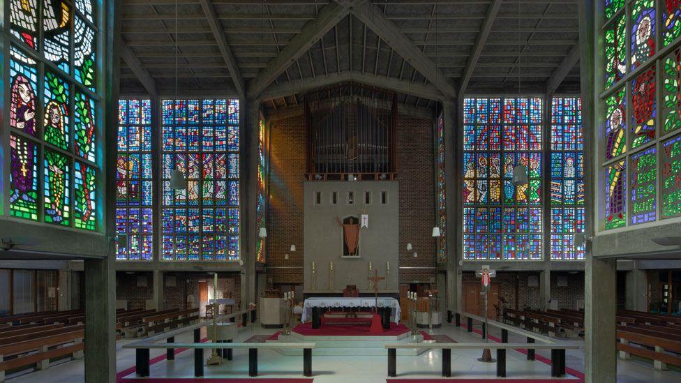 The interior of mid-20th Century Roman Catholic Church of Our Lady of Fatima, Harlow. On the right and left after vividly coloured rectangular stained glass windows raised on concrete legs. In front is an altar with a Christ figure above it and it rises to a concrete beamed roof. On either side of the altar are more huge stained glass rectangular windows