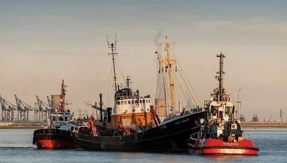 The Arctic Corsair, pictured on water with an industrial skyline in the background. Its colours are black, brown and white. 