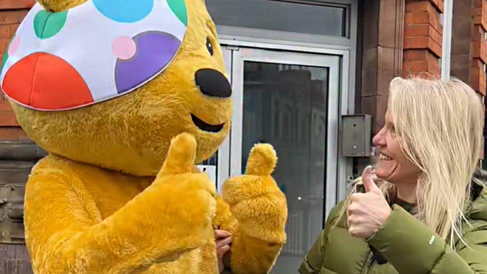 Pudsey bear and Annabel Amos standing outside the BBC Radio Northampton office with their thumbs up, while looking at each other. Pudsey is a yellow bear with a dotty eye-patch. Annabel has long blonde hair and is wearing a green puffer jacket.