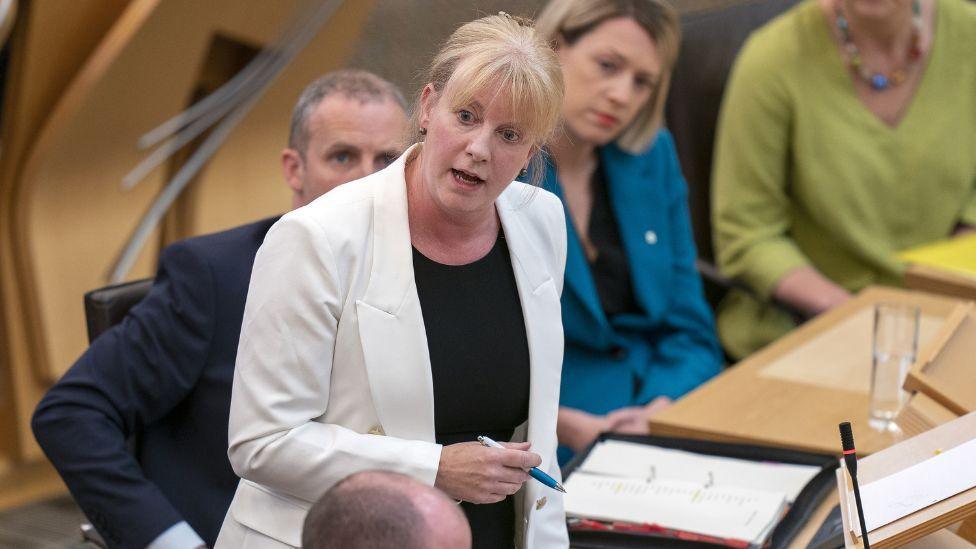 Shona Robison, with light hair and a white jacket, speaking in the Scottish Parliament 