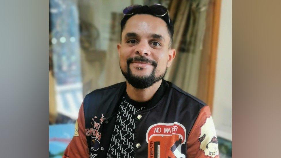 A man with black hair and facial hair. He has sunglasses on his head and is wearing a black and white t-shirt and a leather bomber jacket that is black with orange sleeves and patches all over it.