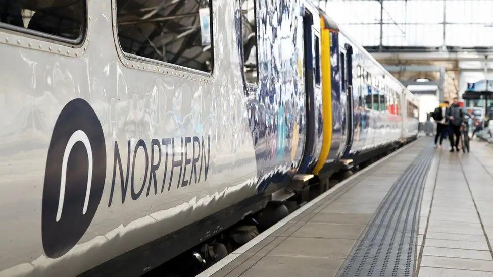 A set of carriages on a Northern train running along a platform with the company sign seen on the outside of one of the cars. People can be seen at the end of platform waiting to alight. 