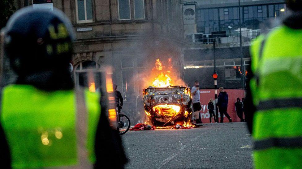 Police in riot gear look on with a burning car in the road in Sunderland