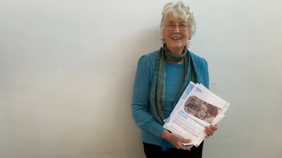 Dee Way standing against a wall smiling and holding a pamphlet