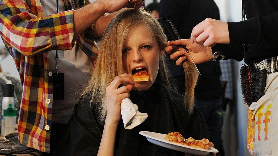 A model takes a bite of pizza while she gets her hair done backstage before the Donna Karan show