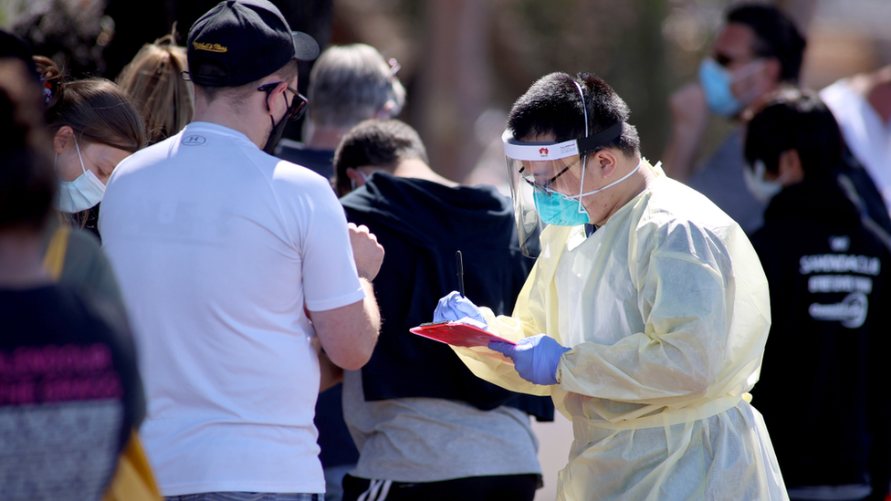 A healthcare worker wearing full protective equipment records details of people queuing to get tested