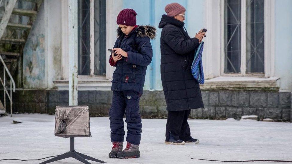 A boy and a woman stand in a snowy scene next to a Starlink terminal, using their electronic devices