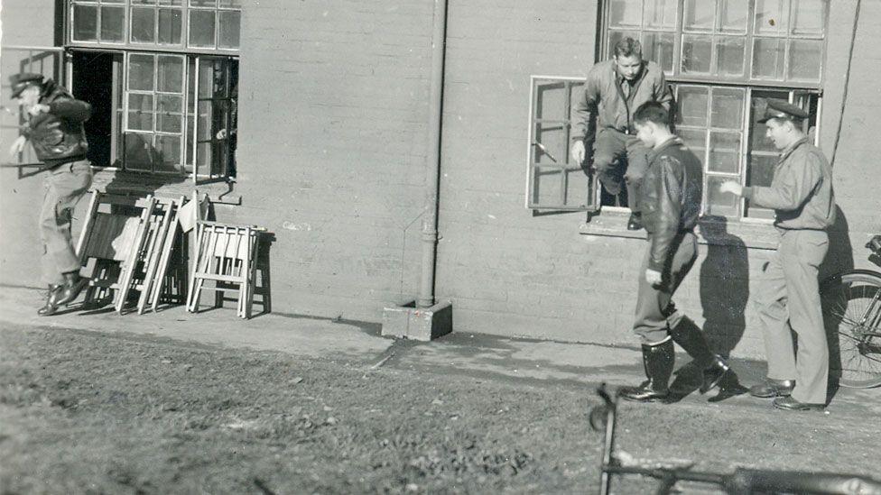A black and white photo of four US airmen at a building with two large windows at RAF Duxford during World War Two. One airman has just jumped out of the  window on the left. On the right, an airman is half in and half out of a window, ready to leap. The two other airman are walking on the pavement ahead of him. 