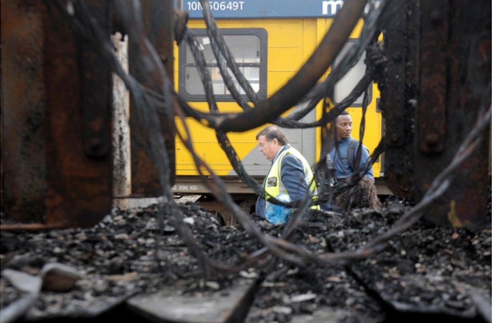 Workers walking along the track next to burnt-out trains are seen through a gap in the wreckage