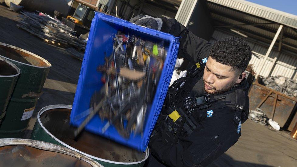 A box full of knives is emptied into a steel drum for crushing