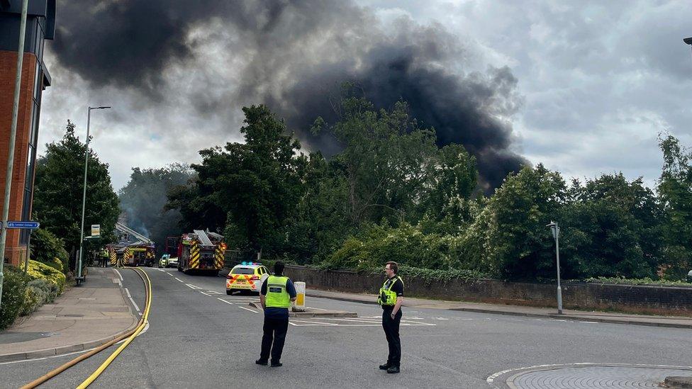 Plume of smoke above car workshop fire, with police officers in foreground, on closed roundabout