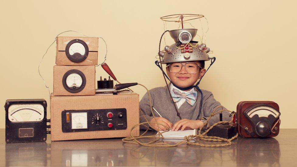 A boy wears a colander on his head and sits next to cardboard boxes connected with string to look like a home made brain scanner.