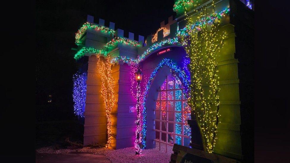 Castle fabrication lit up with bright lights. There are several trees in front of it which each have a different colour of Christmas lights in them. They are yellow, blue, pink and orange. A garland that runs around the edge of the castle has green lights.