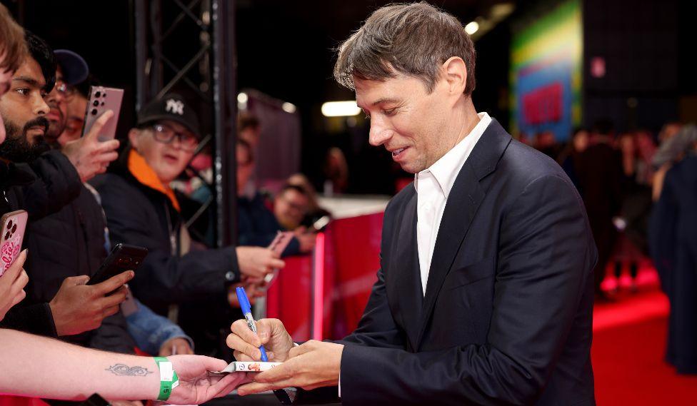 Sean Baker attends the "Anora" Headline Gala during the 68th BFI London Film Festival at The Royal Festival Hall on October 11, 2024 in London, England. He is signing an autograph for a fan while on the red carpet.