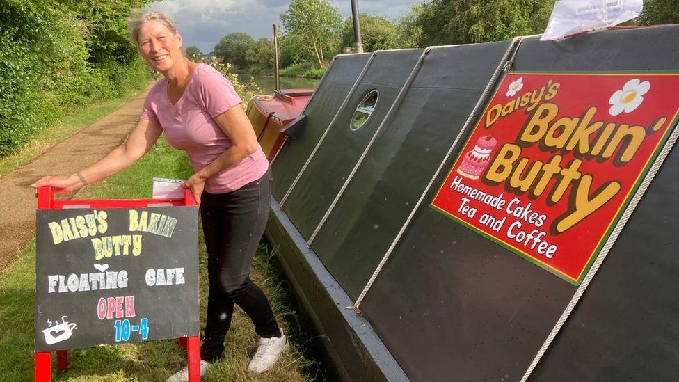 A woman with blonde hair tied back, wearing a pink top and dark leggings, is standing next to her black narrowboat, with a sign saying "Daisy's Bakin' Butty Floating Cafe" which she is putting up on the towpath. The boat also has a red sign with the cafe's name on it. A mud towpath, flanked by a hedge, is on the left.