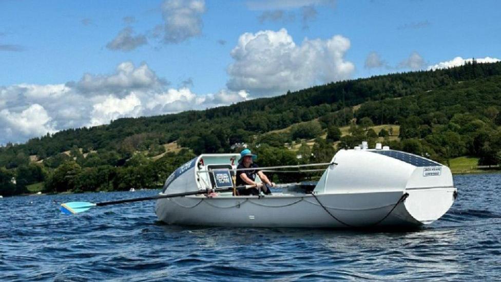 Vivienne Barclay rowing her boat on a lake with hills, trees and green fields behind her.