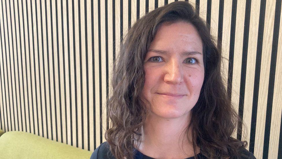 Jessica Rundell who is sitting on a light greeny yellow sofa against a wooden panelled wall. She has dark brown gently curling hair falling to her shoulders and is wearing a dark T-shirt. 