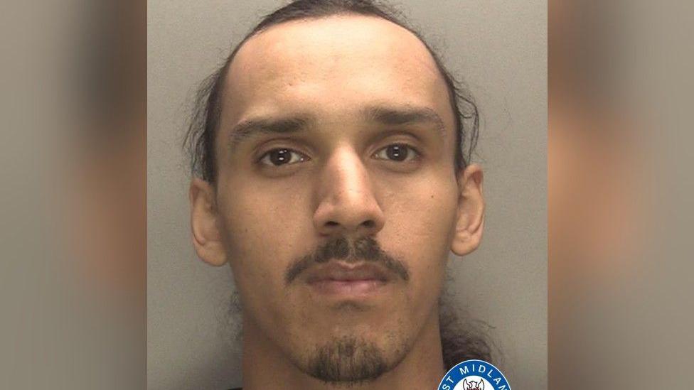 A man with a thin moustache and facial hair on his chin looks at the camera for his mugshot. He is in front of a grey background.