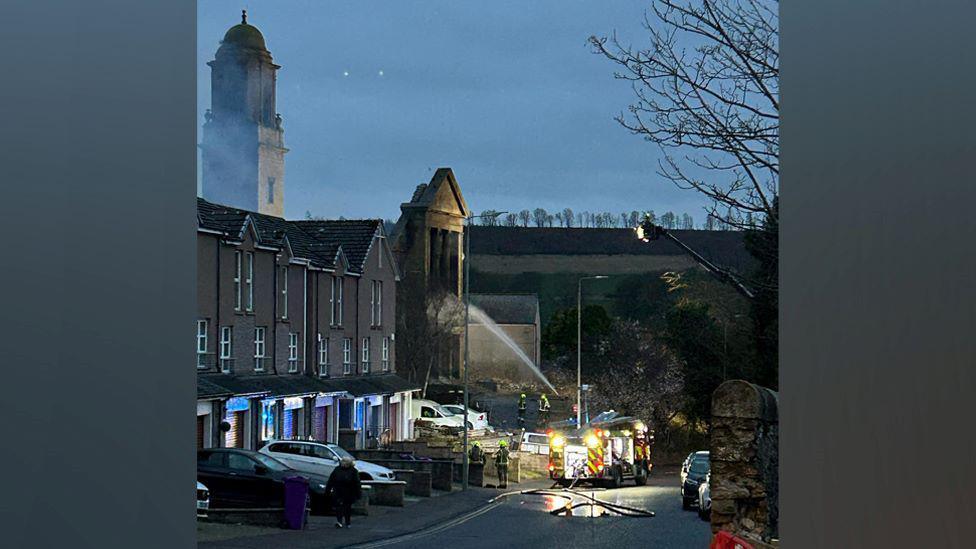 Fire crews battling a fire at a disused church, with a jet of water going into the building, area starting to get dark, fire engine lights can be seen.